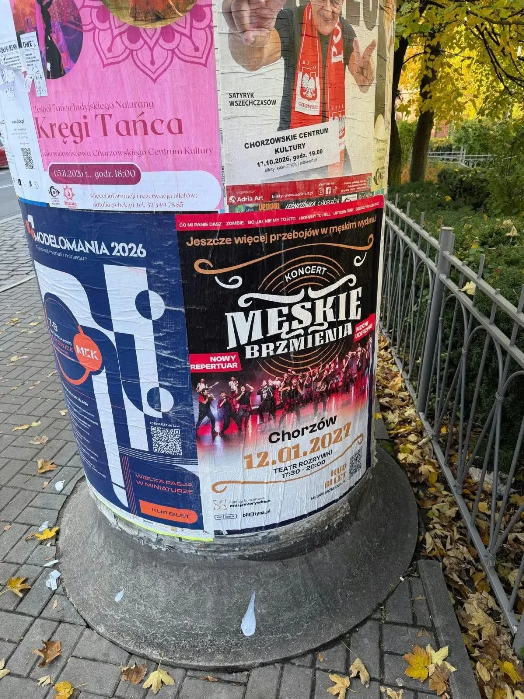 Plakat Męskie Brzmienia na słupie przy metalowym ogrodzeniu i jesiennych liściach