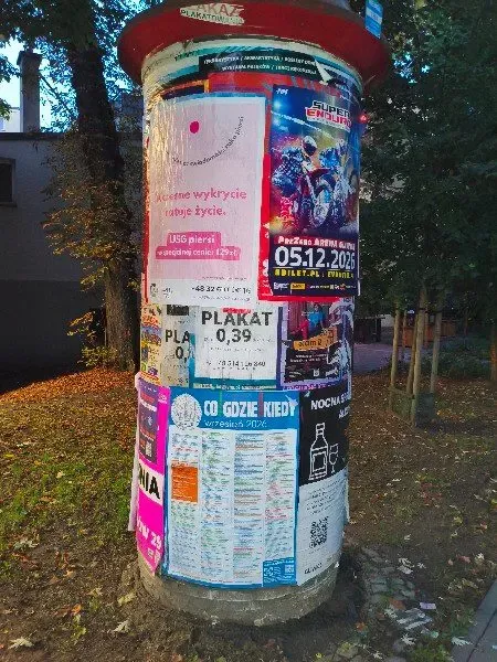 Słup ogłoszeniowy z plakatem USG piersi w cieniu drzew przy chodniku
