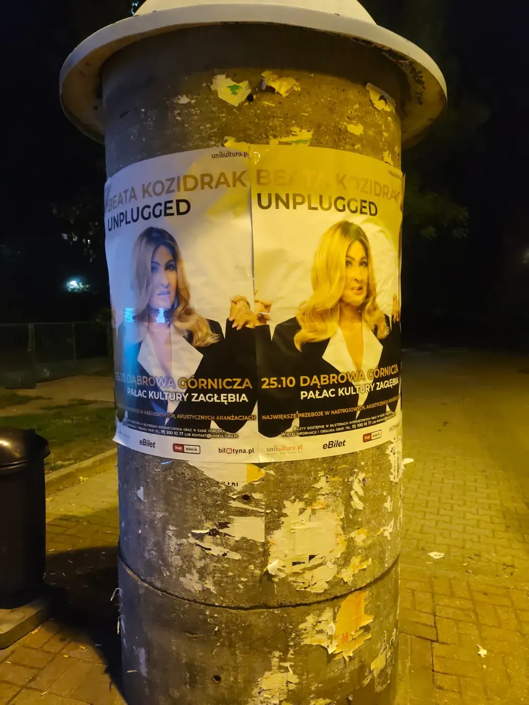 Plakaty koncertowe na słupie ogłoszeniowym przy chodniku w Katowicach