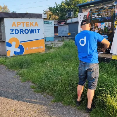 Rozwieszanie plakatów na tablicy ogłoszeń przy osiedlowej ścieżce