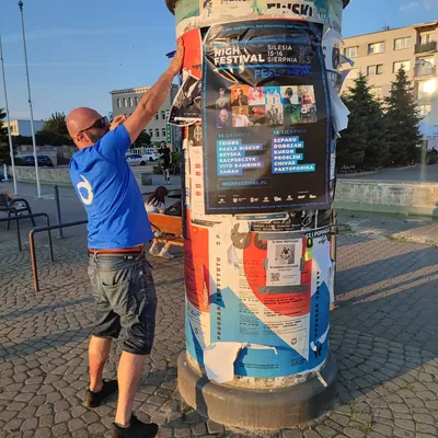 Naklejanie plakatu festiwalu na słupie ogłoszeniowym na placu